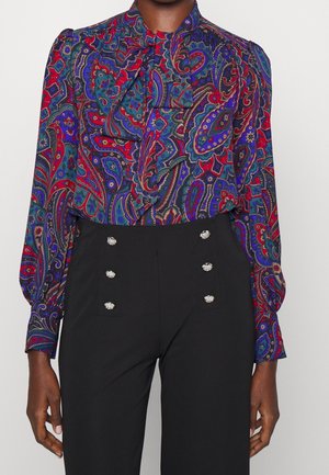 Bluzka z długim rękawem z żywym wzorem paisley w kolorach czerwonym, niebieskim i fioletowym, zestawiona z czarnymi spodniami z wysokim stanem ozdobionymi sześcioma dekoracyjnymi guzikami.