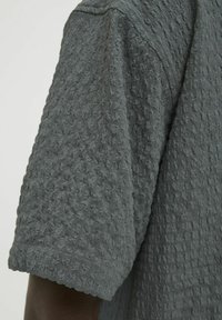 Camisa corta de manga con textura gris, con un patrón de tejido tipo waffle y un escote redondeado, que presenta una superficie de tela suave y elevada.