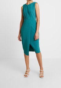 Sininen, hihaton mekko pehmeästä neulospuvusta, jossa on laskostettu muotoilu ja epäsymmetrinen helma, sileä pinta ja minimalistinen tyyli.