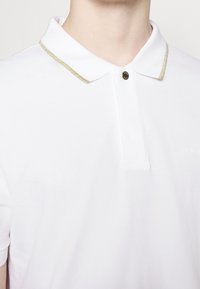 Vit pikétröja i texturerat material, med en guldaccent längs kragen och en svart knapp vid halsliningen.