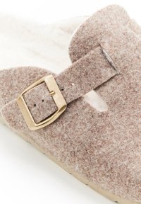 Close-up van een beige vilten sandaal met een gouden rechthoekige gesp op een dikke band over een lichte zool.