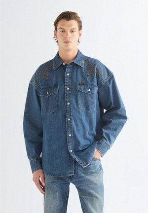 Junger Mann, der ein blaues Jeanshemd mit bestickten Details und blaue Jeans trägt, steht mit einer Hand in der Tasche vor einem schlichten Hintergrund.