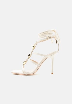 Fehér magas sarkú szandál vékony pántokkal, arany díszítő elemekkel, bokapántos csattal és fonott kötéllel a boka körül.