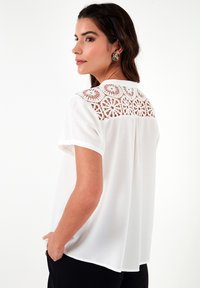 White short-sleeve blouse with a lace panel at the back. Features a relaxed fit and soft texture, complemented by curved hem and subtle detailing.