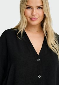 Črna, teksturirana srajca z V-izrezom in štirimi okroglimi gumbi spredaj. Gladka, mehka tekstura in sproščen kroj.