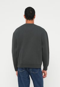 Tmavosivá mikina s okrúhlym výstrihom a dlhými rukávmi, s voľným strihom a rebrovanými lemami, v kombinácii s modrými džínsami.