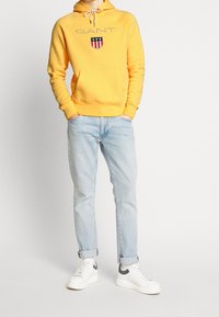 Žlutá mikina s kapucí a přední kapsou s vyšívaným logem, doplněná světle modrými džínami a bílými teniskami s šedými doplňky.