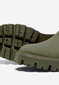 Bottes basses vert olive avec une texture lisse, bout rond et une semelle épaisse avec des nervures pour l'adhérence. Présente un bord cousu en détail.
