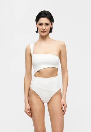 Woman with short dark hair wearing a white asymmetrical one-shoulder cutout swimsuit against a plain light background.