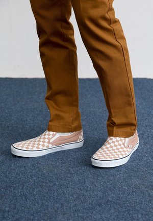 Ben iført brune bukser og lyserøde og hvide ternede slip-on sneakers står på et blåt tæppe.