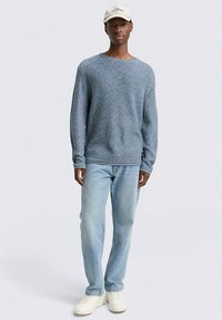 Hellblauer Strickpullover mit rundem Ausschnitt, kombiniert mit hellblauen Jeans und weißen Turnschuhen, getragen von einer stehenden Person.