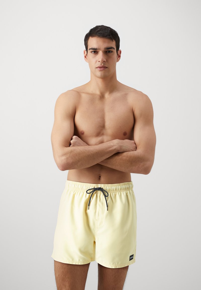 Shorts de bain jaune clair avec une taille élastique et un cordon de serrage noir, ornés d'une petite étiquette de marque noire sur l'ourlet gauche.