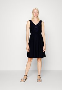 Vestido de marinheiro com design sem mangas, decote em V e detalhe bordado no peito. Cintura com fita e saia fluida. Usado com sandálias leves.