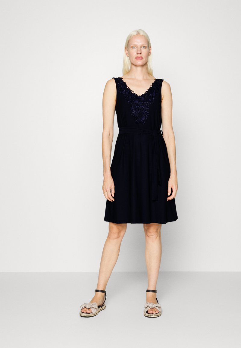 Vestido de marinheiro com design sem mangas, decote em V e detalhe bordado no peito. Cintura com fita e saia fluida. Usado com sandálias leves.