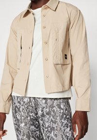 Giacca leggera beige con chiusura a bottoni, due tasche e vestibilità rilassata, indossata sopra una maglietta bianca a coste e pantaloni grigi a motivo camouflage.