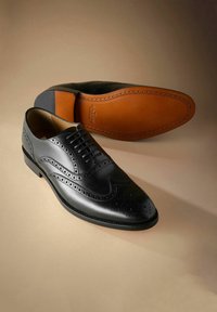Zwarte leren brogue nette schoenen met geperforeerde details, één schoen rechtopstaand en de andere die de beige zool met stiksels toont.
