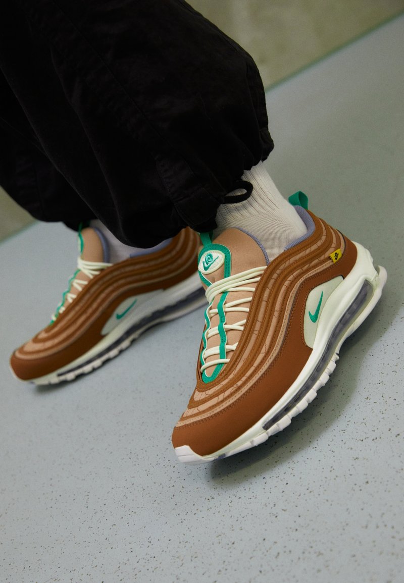 Personne portant des baskets Nike Air Max marron et beige avec des accents bleu sarcelle, des chaussettes blanches et un pantalon noir, debout sur un sol tacheté.