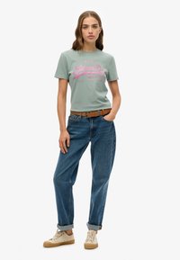 Camiseta de algodón verde claro con estampado gráfico rosa, combinada con unos vaqueros de denim azules holgados y zapatillas beige. Un cinturón marrón acentúa el conjunto.