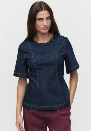 Haut en denim bleu foncé à manches courtes et col rond. Présente des coutures visibles et une silhouette ajustée. Porté avec un pantalon bordeaux.