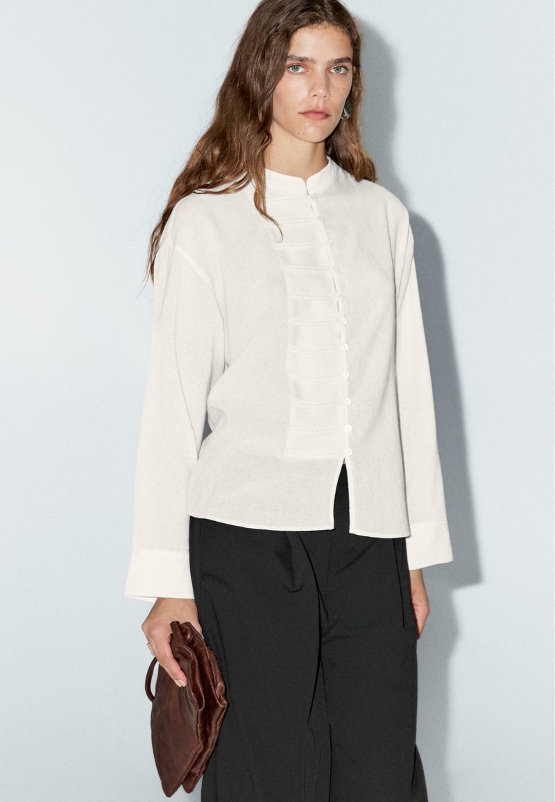 Weißes Hemdblusenshirt mit Stehkragen, ausgestattet mit einem strukturierten Vorderteil und langen Ärmeln. Kombiniert mit schwarzen Hosen und einer braunen Clutch.
