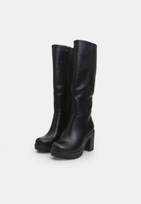 Botas negras hasta la rodilla hechas de cuero liso, con tacón ancho y suela de goma. Detalle de cremallera en el lado interior; diseño elegante y simple.