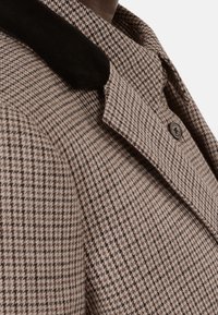 Hnědý kabát s houndstooth vzorem a černým semišovým límcem, s klopovým límcem a kulatým tmavým knoflíkem na přední straně.