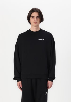 Svart hettegenser med ribbet hals, senkede skuldre og logodetalj på brystet. Myk tekstur, oversized design. Standard passform.