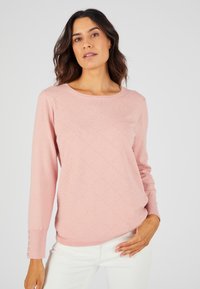 Maglione a maglia rosa chiaro con maniche lunghe, scollatura rotonda, motivo a rombi testurizzato e dettagli con bottoni sui polsini. Indossato sopra pantaloni bianchi.