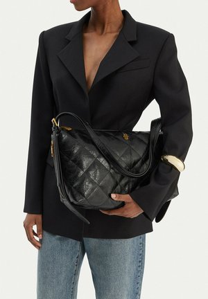 Osoba v černém sakuu a modrých džínách držící velkou černou prošívanou koženou kabelku se zlatými detaily a zlatým náramkem.