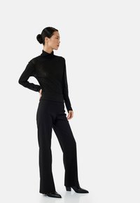 Haut noir à col roulé avec un motif texturé, associé à un pantalon noir taille haute. Le mannequin porte des escarpins noirs à nez pointu, mettant en valeur une silhouette élégante.