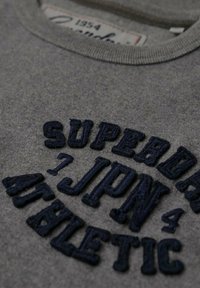 Sudadera de algodón gris con texto bordado en azul oscuro: "SUPERDRY JPN ATHLETIC", con cuello redondo y tejido texturizado.