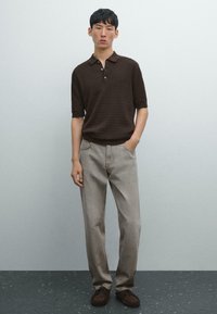 En ung mand står foran en lys grå baggrund, iført en brun polo-shirt, grå jeans og brune slip-ons, med hænderne i lommerne.
