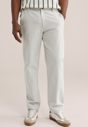 Világosszürke nadrágot, fehér és bézs tornacipőt, valamint csíkos ujjatlan felsőt viselő férfi, kezét a zsebében tartva áll.