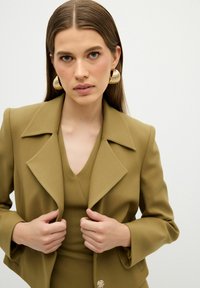 Olijfgroene blazer met een brede revers en een getailleerd silhouet, gecombineerd met een bijpassend top. Gouden statementoorbellen zorgen voor contrast.