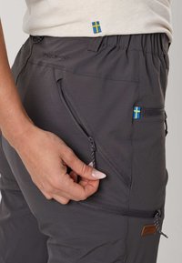 Hand adjusting zipper on dark grey SwedenMount pants with Swedish flag tags and elastic waistband, wearing light beige top.