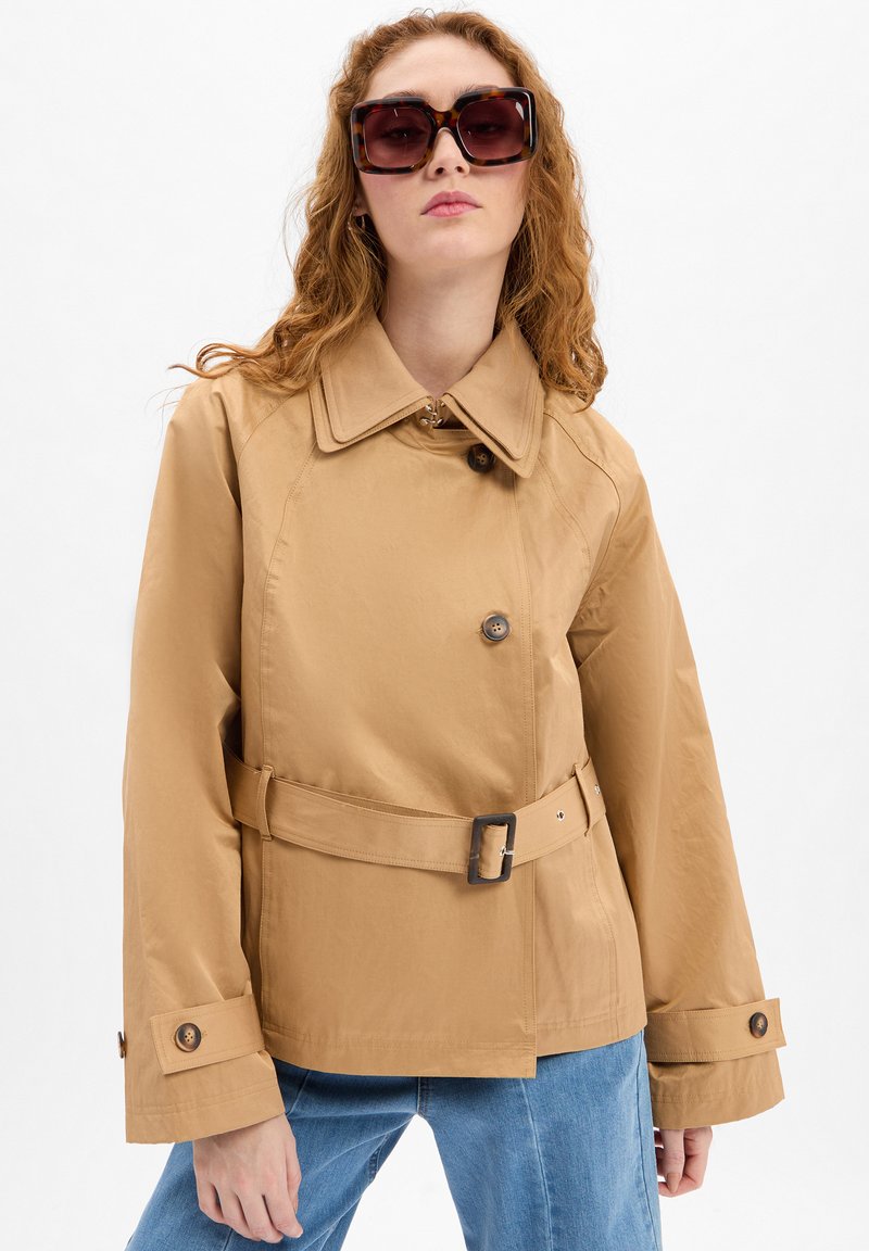 Tan trenchcoat met een riem in de taille, grote kraag en knoopdetails. G gedragen met oversized, donkere schildpadvormige zonnebril.