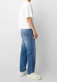 Camiseta blanca de algodón de manga corta, combinada con unos jeans de mezclilla azul claro de corte recto y bolsillos traseros clásicos. Zapatillas deportivas de color blanco sucio.