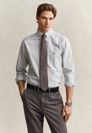 Homme portant une chemise grise rayée à boutons avec manches retroussées, cravate à motifs, pantalon gris, ceinture noire et montre-bracelet argentée, debout les mains dans les poches.