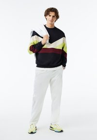 Sudadera navy con rayas en forma de chevron en verde, crema y marrón. Pantalones blancos y zapatillas amarillas con acentos en negro. Modelo sosteniendo una chaqueta blanca.