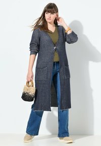 Nainen pukeutunut pitkään tummanharmaaseen neulekarvahintaan, jonka alla on oliivinvihreä toppi ja siniset farkut, pitelee kaksiväristä viinipullokassia, jossa on puukahvat.