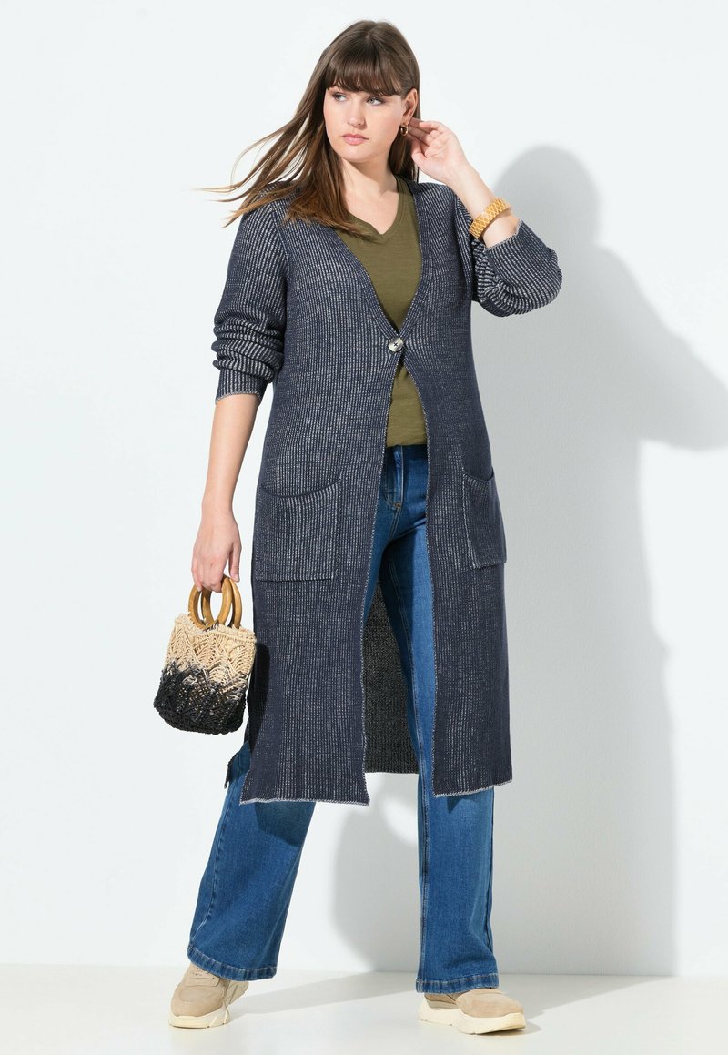 Nainen pukeutunut pitkään tummanharmaaseen neulekarvahintaan, jonka alla on oliivinvihreä toppi ja siniset farkut, pitelee kaksiväristä viinipullokassia, jossa on puukahvat.