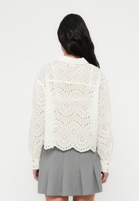 Chemise blanche brodée avec un motif ajouré, manches longues, ourlet festonné et détails de boutons. Portée par-dessus une jupe grise.