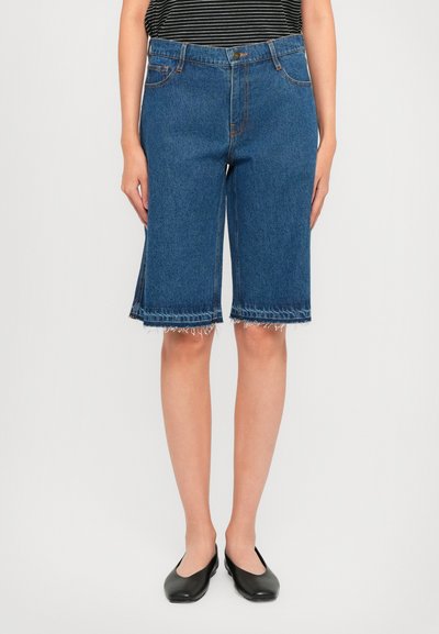 Vrouw in knielange blauwe denim shorts met rafelige zomen, zwarte flats en een zwart-wit gestreept T-shirt tegen een effen witte achtergrond.