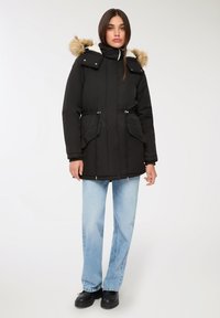 Czarna ocieplana parka z kapturem wyłożonym futrem, z pozwalającą na regulację talią i dwoma przednimi kieszeniami. Połączona z jasnoniebieskimi prostymi dżinsami i czarnymi butami.