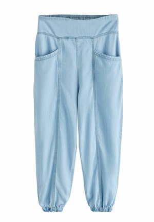 Personne portant un pantalon gris clair à taille élastique et une chemise bleue, soulevant légèrement la chemise pour dévoiler la ceinture à l'extérieur.