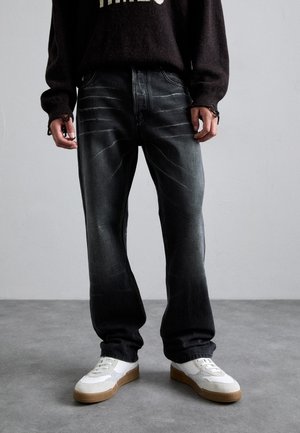 Jean en denim noir avec une finition délavée, coupe droite et détails de couture visibles. Associé à des baskets blanches avec une semelle en caoutchouc.
