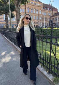 Svart kappa över en vit polotröja, mörka vida byxor och svarta ankelstövlar; solglasögon. Bakgrunden visar en tegelbyggnad och grönska.