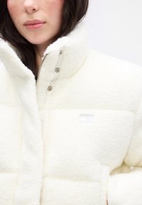 Biely, nadýchaný flísový kabát s vysokým golierom, zipsom a zacvakávacími uzávermi, s jemnou nášivkou loga na ľavej strane.