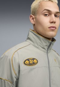 Lysegrå zip-jake med høj krave, gul syning og broderet lilla og gule Sonic the Hedgehog-patch på brystet.