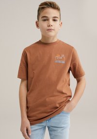 Ruda medvilninė marškinėliai su trumpomis rankovėmis, su mėlynos ir oranžinės spalvos grafikiniu logotipu, kuriame parašyta "BLUE RIDGE DENIM" kairėje krūtinės pusėje.